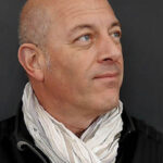 A photo Freddy Silva a man with a bald head and light complexion, wearing a white scarf and a black jacket. He is looking slightly to the right, against a plain dark grey background.