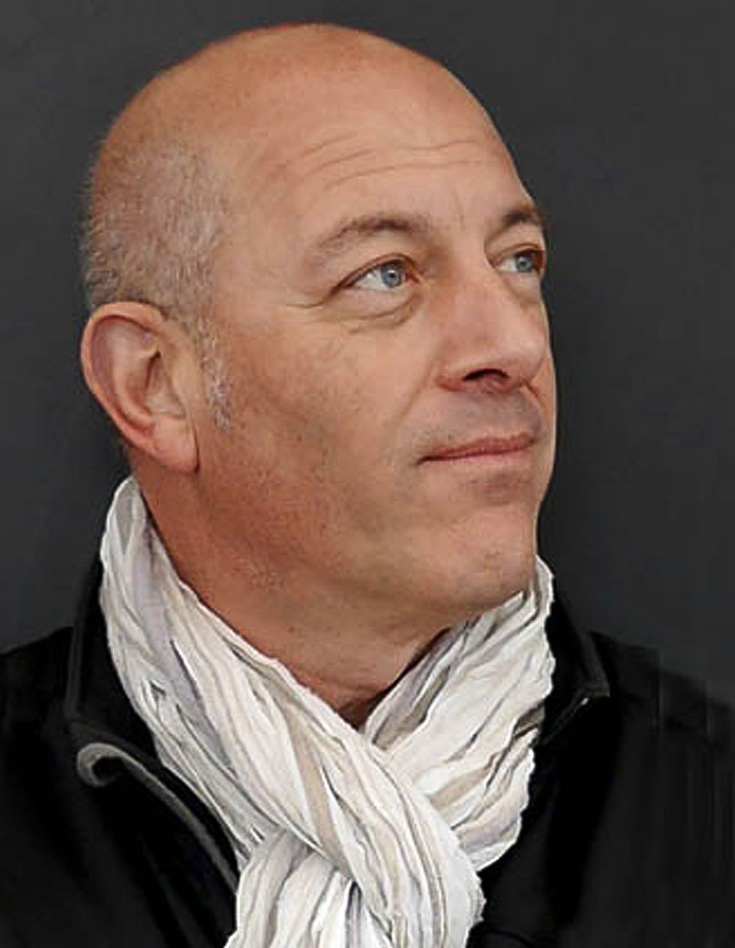 A photo Freddy Silva a man with a bald head and light complexion, wearing a white scarf and a black jacket. He is looking slightly to the right, against a plain dark grey background.