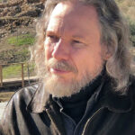 A photo of Dr. Robert Schoch, an individual with long, wavy gray hair and a beard. He is wearing a black leather jacket and a black shirt. The background features the archaeological site of Göbekli Tepe.