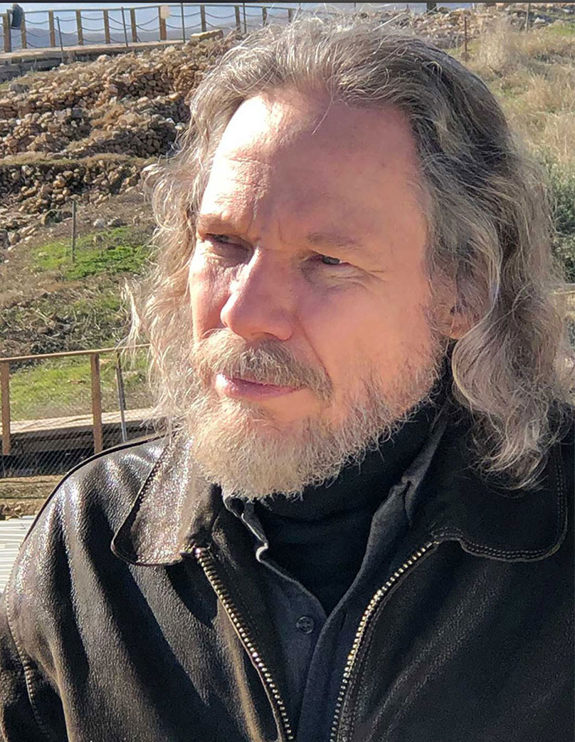 A photo of Dr. Robert Schoch, an individual with long, wavy gray hair and a beard. He is wearing a black leather jacket and a black shirt. The background features the archaeological site of Göbekli Tepe.